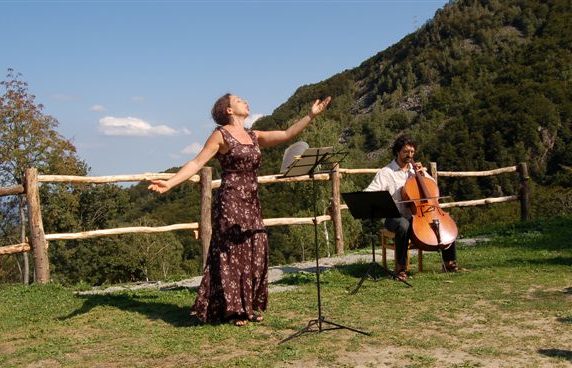 Figura intera della protagonista che si esibisce, davanti a lei un leggio. Alla sua destra un musicista suona un violoncello.