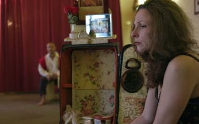 Helena, in primo piano girata di lato, guarda nel vuoto con sguardo triste e preoccupato. In lontananza, sfocato, tra l'arredamento, si scorge Giorgio.