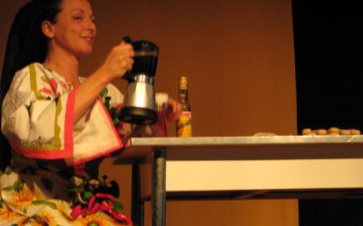 La protagonista, vestita di panni da cucina, tovaglie coi girasoli e peperoncini, è seduta al tavolo e si versa una tazza di caffè.