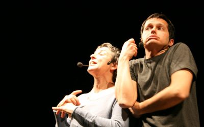 Piano americano dei due attori in scena, sfondo nero. Lei a sinistra e lui a destra, sono molto vicini. Entrambi gesticolano e sono in movimento.
