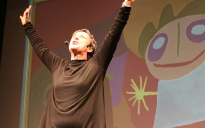 Piano americano dell'attrice sul palco, parla con il pubblico e alza le mani al cielo. Dietro di lei un disegno proiettato, coloratissimo, raffigurante delle persone stilizzate.