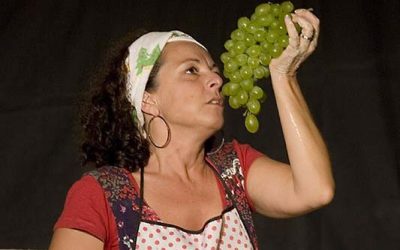 Primo piano della protagonista con in mano un grappolo d'uva bianca, che guarda, tenuto in alto vicino alla bocca.