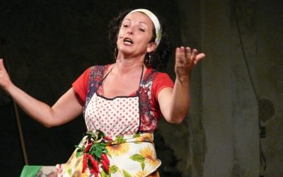 Primo piano della protagonista, vestita con una tovaglia da tavolo con i girasoli per gonna, un grembiule da cucina e una maglietta rossa. Ha in vita un grappolo peperoncini.