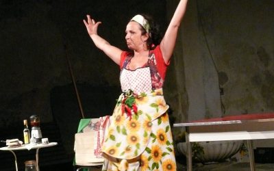 La protagonista è in piedi sul palco. è vestita con una tovaglia da tavolo con i girasoli per gonna, un grembiule da cucina e una maglietta rossa. Ha in vita un grappolo di peperoncini.
