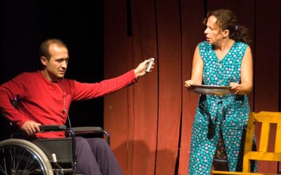 I due protagonisti in scena. Giorgio, sulla sedia a rotelle, con un telecomando in mano, a sinistra, e Helena, con un vassoio in mano, a destra.