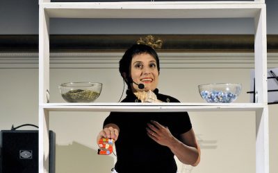 Piano americano di un'attrice, dietro uno scaffale bianco in cui sono poggiate due ciotole piene di pietre colorate verdi e azzurre e una conchiglia. L'attrice tiene in mano e mostra al pubblico una S rossa con dei fiorellini.