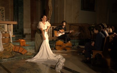 La lavandaia del Presepe raccoglie da terra le fasce del Bambinello. Dietro di lei un musicista suona la chitarra. A destra si vede la prima fila del pubblico.