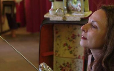 In primo piano, Helena ascolta una radiolina, che tiene in mano, con sguardo trasognato. In lontananza, sfocato, tra l'arredamento, si scorge Giorgio.